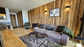 A modern living room with a wooden wall panel, a grey sofa, two round coffee tables, and a wooden side table. - Appartement T2-Cabine B-02 - Collection Cassiopée (Font-Romeu-Odeillo-Via)