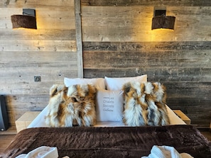 A bed with two fluffy pillows and a white pillow with text, set against a wooden wall with two wall-mounted lights.