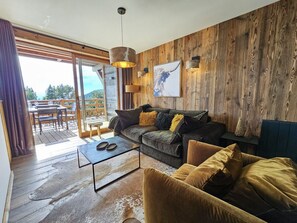A living room with a wooden wall, a sofa, a coffee table, and a view through a glass door to an outdoor area. - Appartement A-12 T2 Cabine (Font-Romeu-Odeillo-Via)