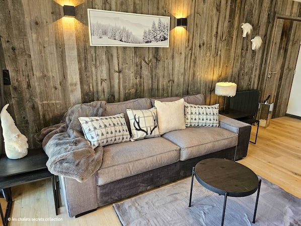Uma sala de estar acolhedora com uma parede de madeira, um sofá com almofadas estampadas, uma mesa lateral e uma pequena mesa redonda.