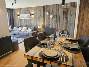 Uma sala de jantar com mesa de madeira posta para quatro pessoas, rodeada por cadeiras. Um sofá e um radiador são visíveis ao fundo.