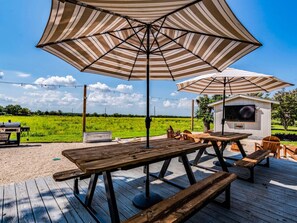 Eine Außenpatio mit Picknicktischen und gestreiften Sonnenschirmen, ein Grill und ein Fernseher in einem kleinen Gebäude.