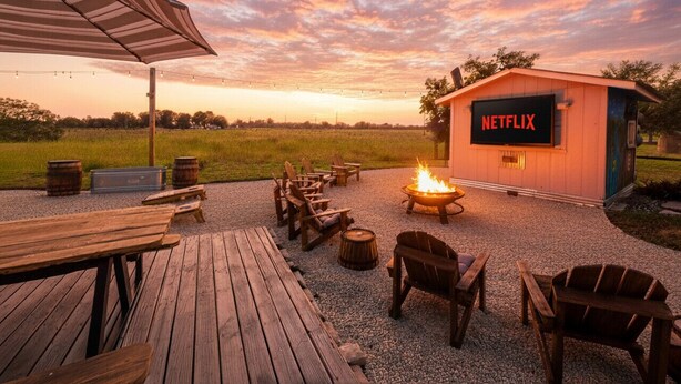 An outdoor seating area with wooden tables and chairs, a fire pit, and a building labeled "NETFLIX" in the background. - Riverbend Hideaway - 5 Min to Downtown & Tubing (San Marcos)