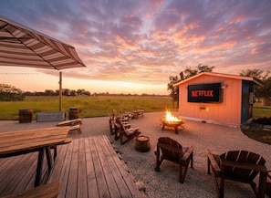 An outdoor seating area with wooden tables and chairs, a fire pit, and a building labeled "NETFLIX" in the background. - Riverbend Hideaway - 5 Min to Downtown & Tubing (San Marcos)