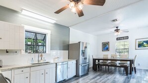A modern kitchen with white cabinets, a stainless steel refrigerator, and a dining area with a wooden table and chairs. - Riverbend Hideaway - 5 Min to Downtown & Tubing (San Marcos)