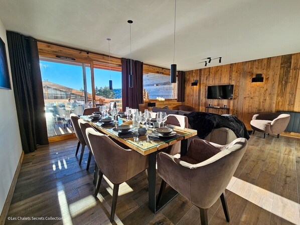 A modern dining area with a glass table, wooden walls, and a large window offering a view of the outdoors. - Appartement A-11 T4 (Font-Romeu-Odeillo-Via)