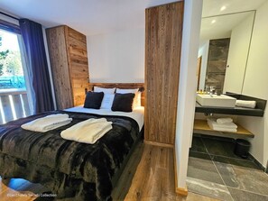 A modern bedroom with a wooden headboard, a large bed with black and white bedding, and a bathroom with a dark tiled floor and a white sink. - Appartement  A-11  T4 (Font-Romeu-Odeillo-Via)