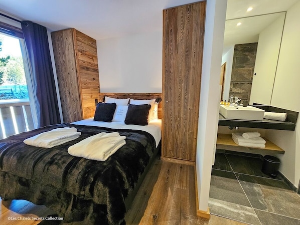 Un dormitorio moderno con cabecera de madera, una cama grande con ropa de cama blanco y negro, y un baño con piso de baldosas oscuras y un lavamanos blanco.