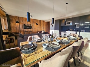 Un comedor moderno con una larga mesa de madera dispuesta para una comida, una cocina al fondo y un rincón de estar acogedor con un sofá y sillas.