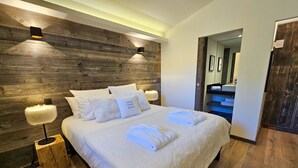 Une chambre moderne avec une tête de lit en bois, un grand lit recouvert de linge de maison blanc, deux lampes de chevet et une salle de bain visible au fond.