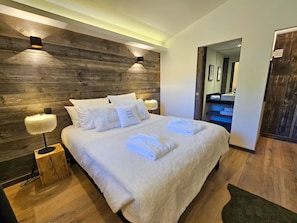 A modern bedroom with a wooden headboard, a large bed with white linens, two bedside lamps, and a visible bathroom in the background.