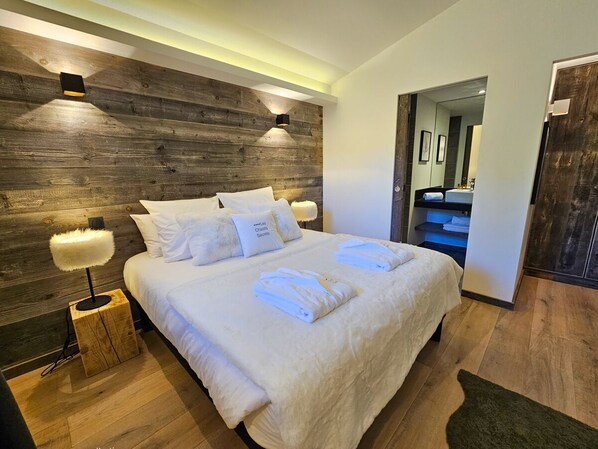 A modern bedroom with a wooden headboard, a large bed with white linens, two bedside lamps, and a visible bathroom in the background.