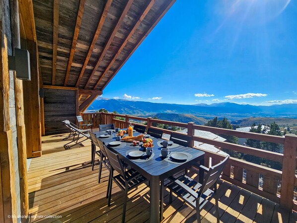 Um deck de madeira com uma mesa posta para o café da manhã, com vista para uma paisagem montanhosa.