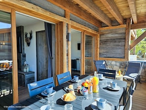 A wooden deck with a table set for breakfast, including croissants and fruit, and a stone fireplace in the background. - Appartement T2-CABINE A-02 - Collection Cassiopée (Font-Romeu-Odeillo-Via)
