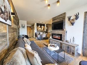 A modern living room with a fireplace, a large TV, and a dining area. - Appartement T2-Cabine A-02 - Collection Cassiopée (Font-Romeu-Odeillo-Via)