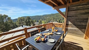An outdoor dining area with a table set for two, surrounded by wooden railings, overlooking a forested landscape.