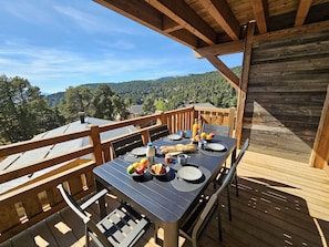 An outdoor dining area with a table set for two, surrounded by wooden railings, overlooking a forested landscape.