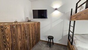 A room with a bunk bed, a wooden wardrobe, a small stool, a television mounted on the wall, and a fluffy rug on the floor.