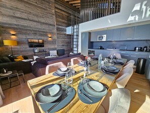 Uma sala de jantar moderna com uma mesa de madeira posta para refeição, um sofá e uma cozinha ao fundo.