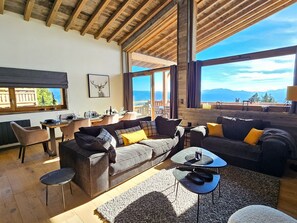 A spacious living area with a large sofa, a coffee table, and a dining table. There is a window with a view of mountains and a wooden ceiling.