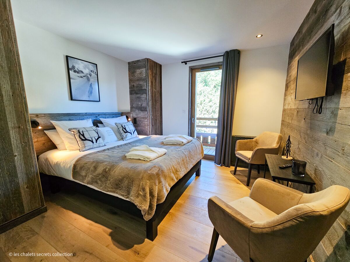 Une chambre avec un grand lit, un bureau et une chaise. Il y a une fenêtre avec des rideaux, un téléviseur fixé au mur et un tableau encadré accroché au mur.