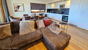 A modern living room with a sectional sofa, a dining area, and a kitchen with white cabinets and stainless steel appliances.