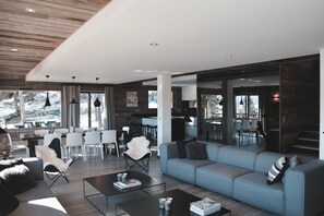 Un salón moderno con sofá gris, sillas blancas y una mesa de centro. Hay una zona de cocina con una mesa de comedor y sillas. Grandes ventanales ofrecen vistas al exterior.