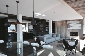 Una sala moderna con un sofá gris, sillas negras y una mesa de centro de vidrio. Se observan luces colgantes y una chimenea.