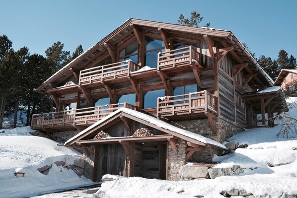 A multi-level wooden cabin with balconies, stone foundation, and snow-covered surroundings.