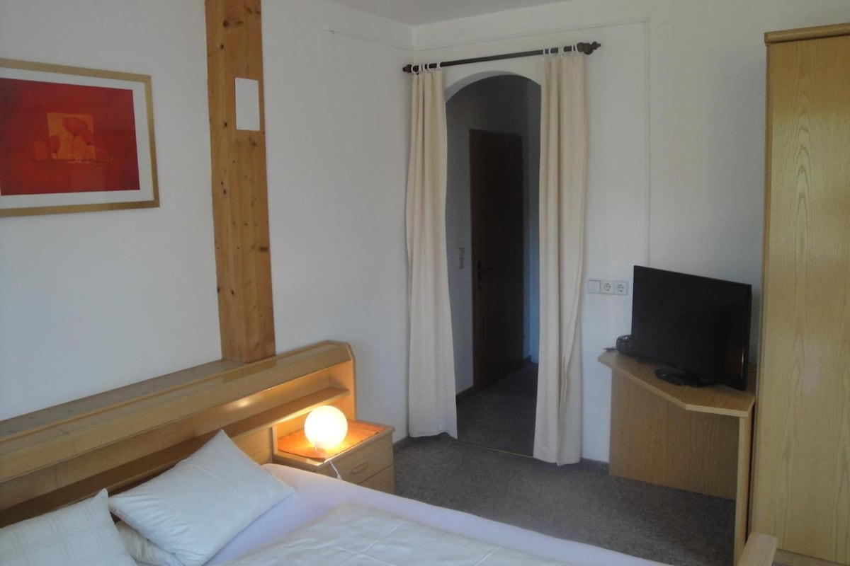 A hotel room with a bed, a bedside lamp, a television on a wooden stand, a door with a curtain, and a framed picture on the wall.