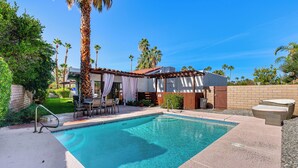 A rectangular swimming pool with clear blue water, surrounded by a patio area with outdoor furniture, a pergola with white curtains, and palm trees in the background.
