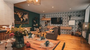 A modern living room with a wooden dining table set for four, a sofa, a TV mounted on the wall, and a patterned wallpaper. - Maison de Campagne 4 Avec Terrain de Pétanque, Idéal Pour les Familles et Amis (Vireux-Wallerand)
