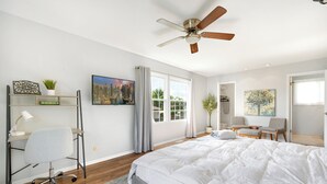 A modern bedroom with a large bed, a desk with a chair, a ceiling fan, and a view of the outdoors through a window.
