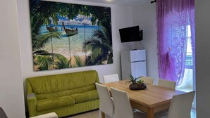 A room with a green sofa, a wooden dining table with chairs, a flat-screen TV mounted on the wall, and a large tropical beach painting.