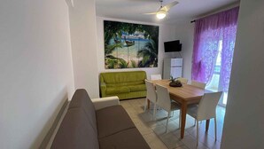 A living room with a sofa, a dining table with chairs, a TV on the wall, and a tropical mural.