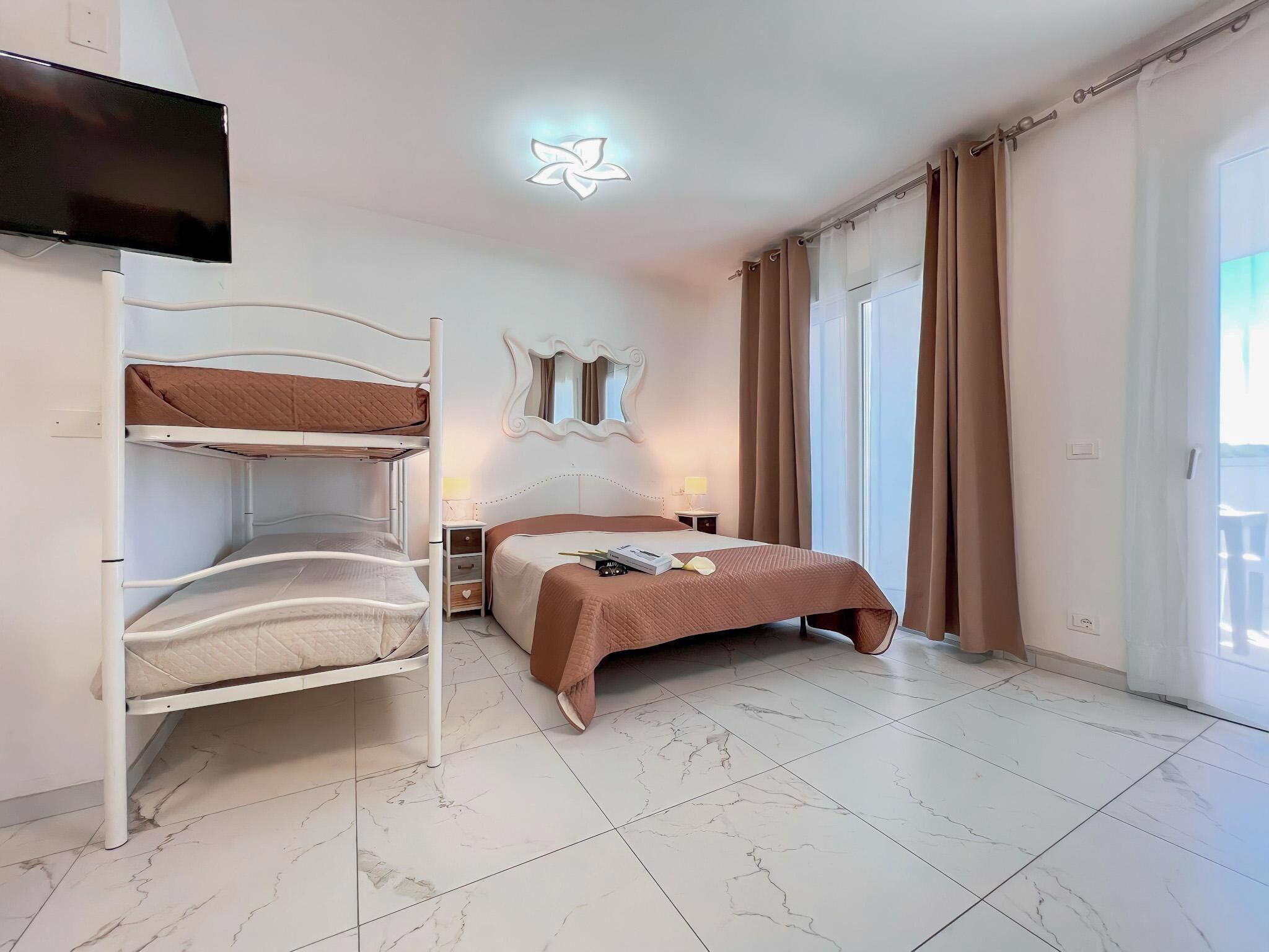A hotel room with a bunk bed, a single bed, a TV, a mirror, and a balcony with curtains.