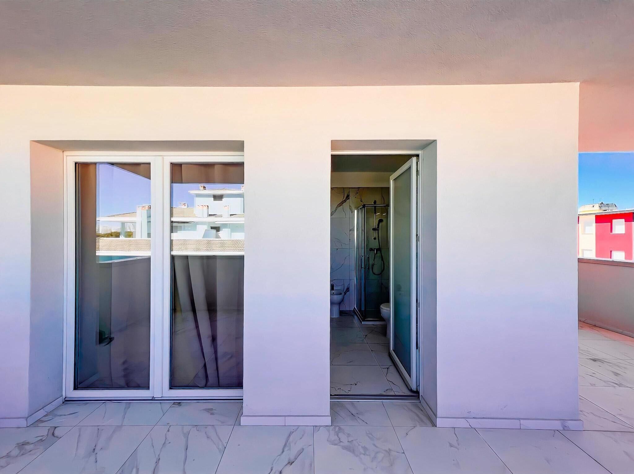 A modern apartment with a glass door leading to a balcony, a shower room, and a view of neighboring buildings.