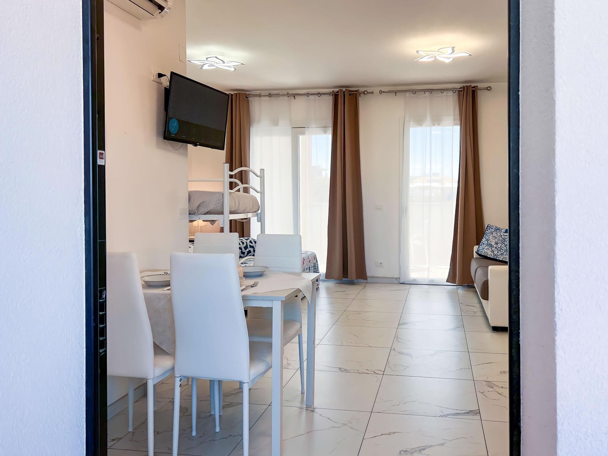 Camera d'albergo moderna con zona pranzo, TV a parete, letto a castello e una porta vetrata scorrevole che dà su un balcone.