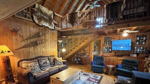 Interior rústico de una cabaña con una escalera de madera, un sofá con cojines de motivos animales, un televisor y un televisor de gran tamaño montado en la pared mostrando una foto grupal.
