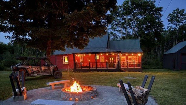 Un espacio de descanso al aire libre de estilo rústico con un hogar, bancos y un edificio rojo al fondo.