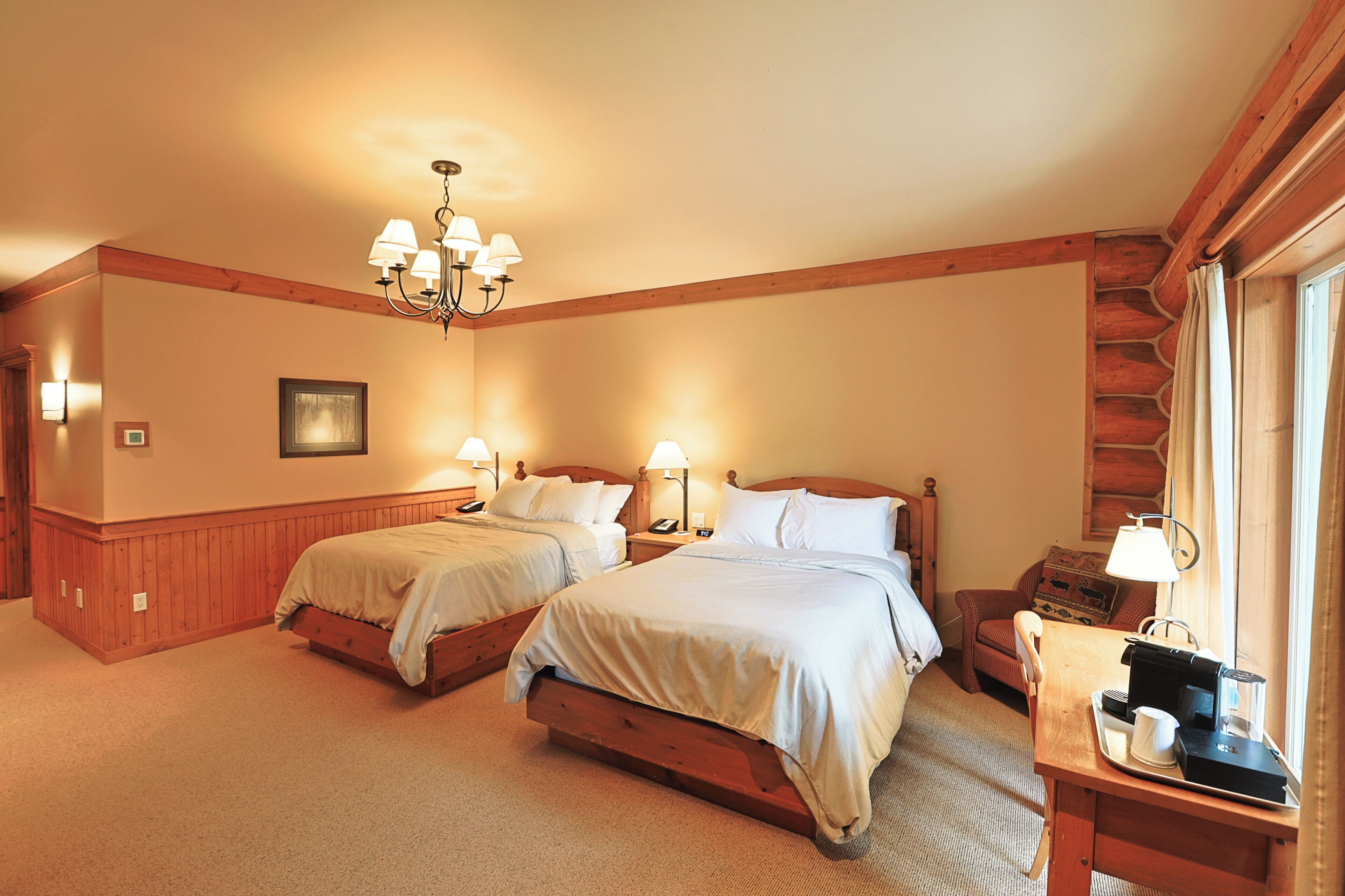 A hotel room with two beds, a wooden desk with a lamp, a chair, and a window with curtains.