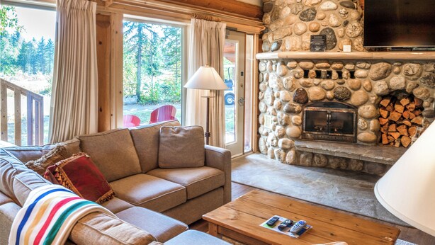 A cozy living room with a stone fireplace, wooden ceiling, and a sofa with a striped blanket. - Mike Wiegele Helicopter Skiing (Blue River)