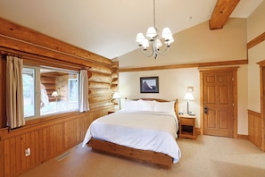 A log cabin bedroom with a large bed, wooden walls, a chandelier, a window with curtains, and a door. - Mike Wiegele Helicopter Skiing (Blue River)