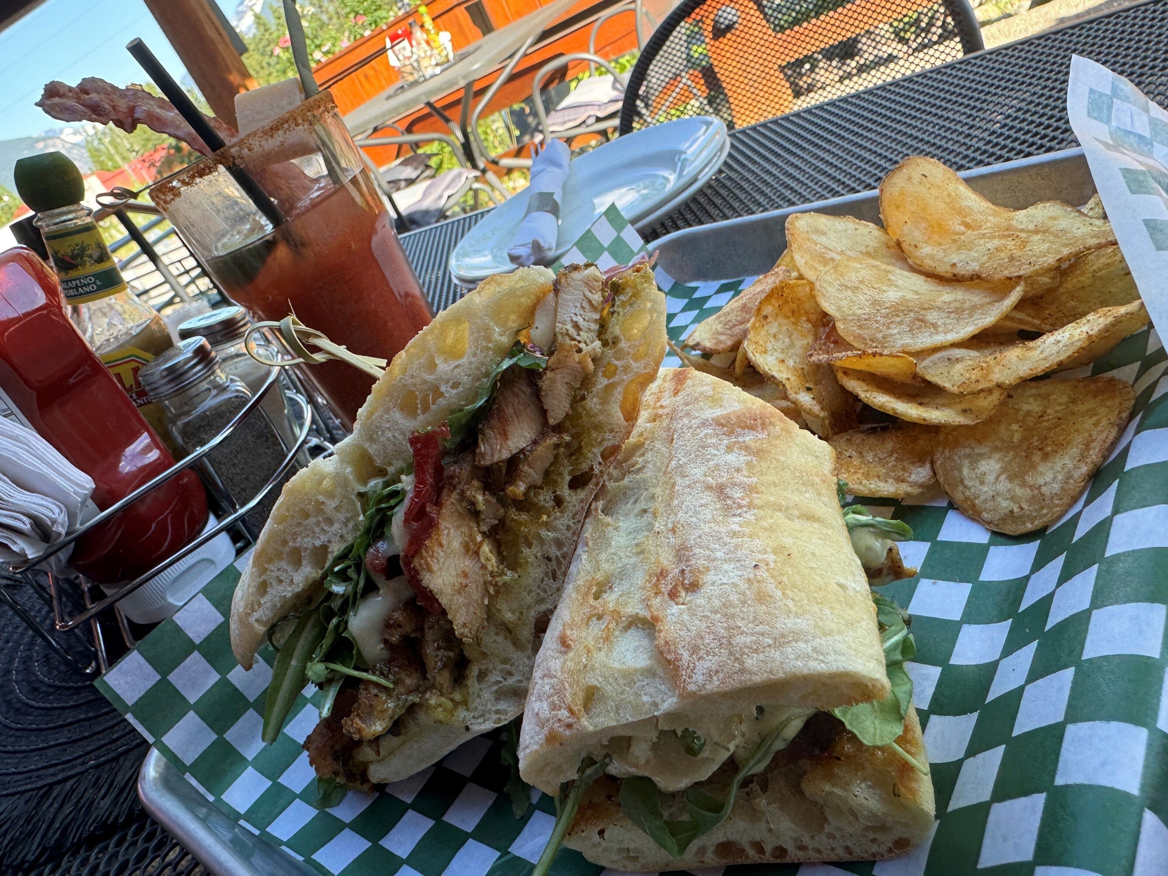 A sandwich on a checkered paper, a bottle of ketchup, a bottle of hot sauce, and a basket of chips.