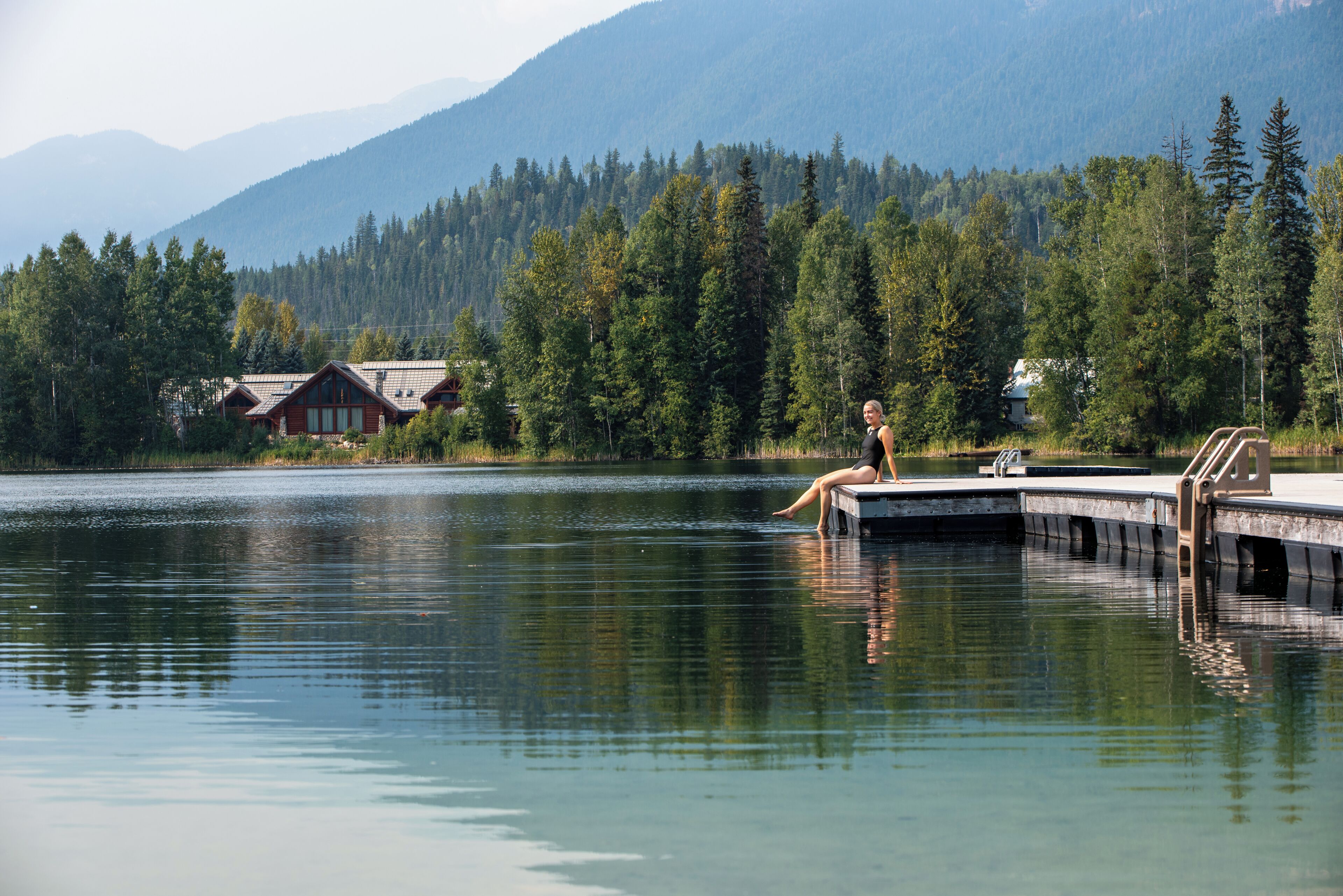 A lakeside cabin with a dock extending into the water, surrounded by dense forest and mountains in the background.