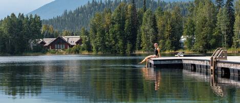 A lakeside cabin with a dock extending into the water, surrounded by dense forest and mountains in the background.