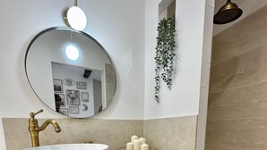 A bathroom with a round mirror, a white sink, a gold faucet, and a showerhead. There are woven baskets under the sink and a plant hanging on the wall. - Cocon Cosy & Chaleureux à Pézenas: Chez Roméo (Pézenas)