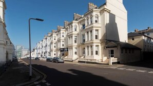 Una hilera de casas urbanas blancas y clásicas, con arquitectura uniforme, cielo despejado y una calle con coches estacionados.