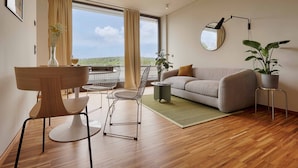 A modern living room with a sofa, a dining table with chairs, a potted plant, and a view through large windows.