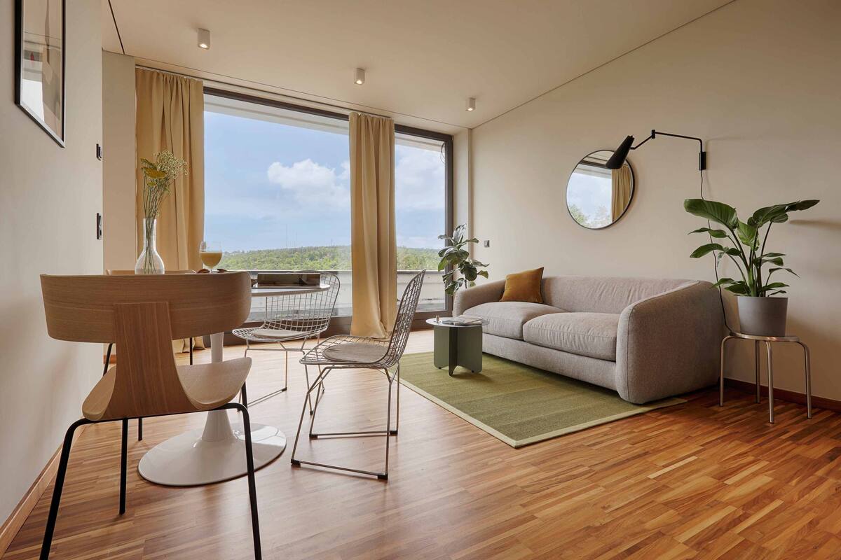 A modern living room with a sofa, a dining table with chairs, a potted plant, and a view through large windows.